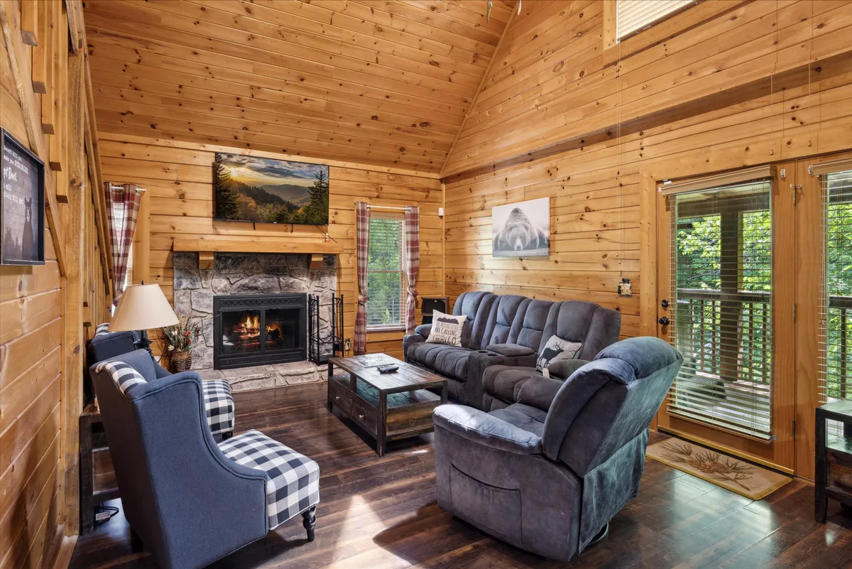 A warm and welcoming living room featuring a stone fireplace as its centerpiece, cozy seating arranged for conversation, and expansive windows that flood the room with natural light. Mountain landscape artwork hangs above the fireplace, and the windows offer a glimpse of the lush surroundings outside.