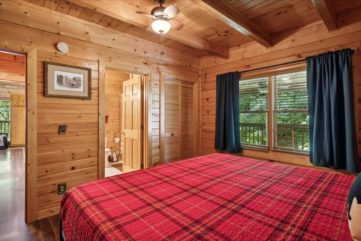 A warmly lit bedroom featuring a plush red plaid bedspread, rustic wooden ceiling, a large window framed with curtains, and cozy framed artwork. The room exudes comfort and has tranquil views of nature, ideal for restful relaxation.