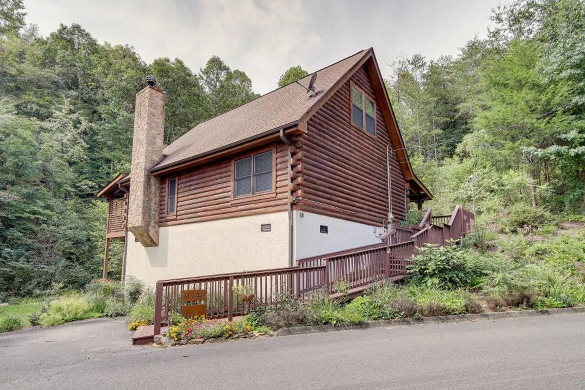 A full view of the log cabin's exterior, with a prominent stone chimney, a spacious wooden deck, and lush greenery all around. The scene is tranquil and welcoming, suggesting a place where families can gather outdoors, enjoy nature walks, and relax together.