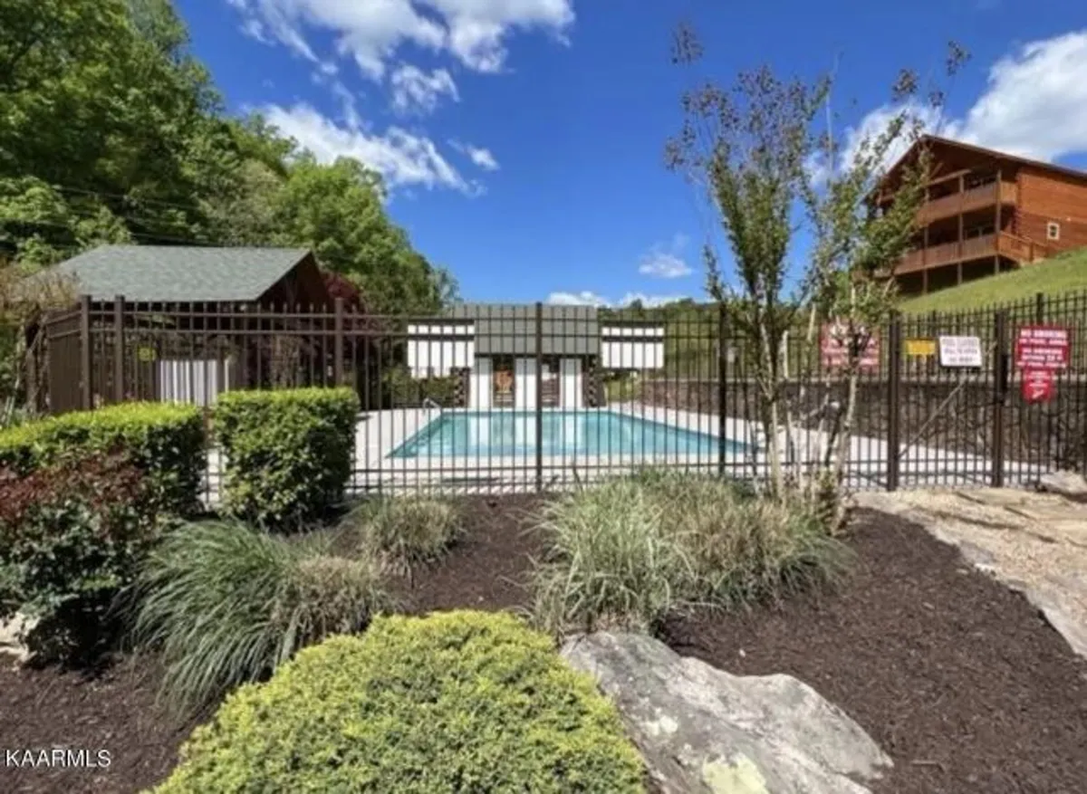 A sparkling swimming pool is the centerpiece, enclosed by a fence for safety, with surrounding greenery and a rustic wooden structure in the background. The pool area is well-maintained, inviting, and clearly designed for community use, suggesting both relaxation and social opportunities.