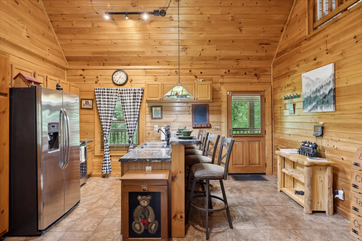 This image highlights a kitchen area with stainless steel refrigerator, wooden cabinetry, bar stools, a large window with a view of surrounding greenery, and decorative wall art. The kitchen is spacious and designed for both cooking and entertaining, with plenty of natural light.