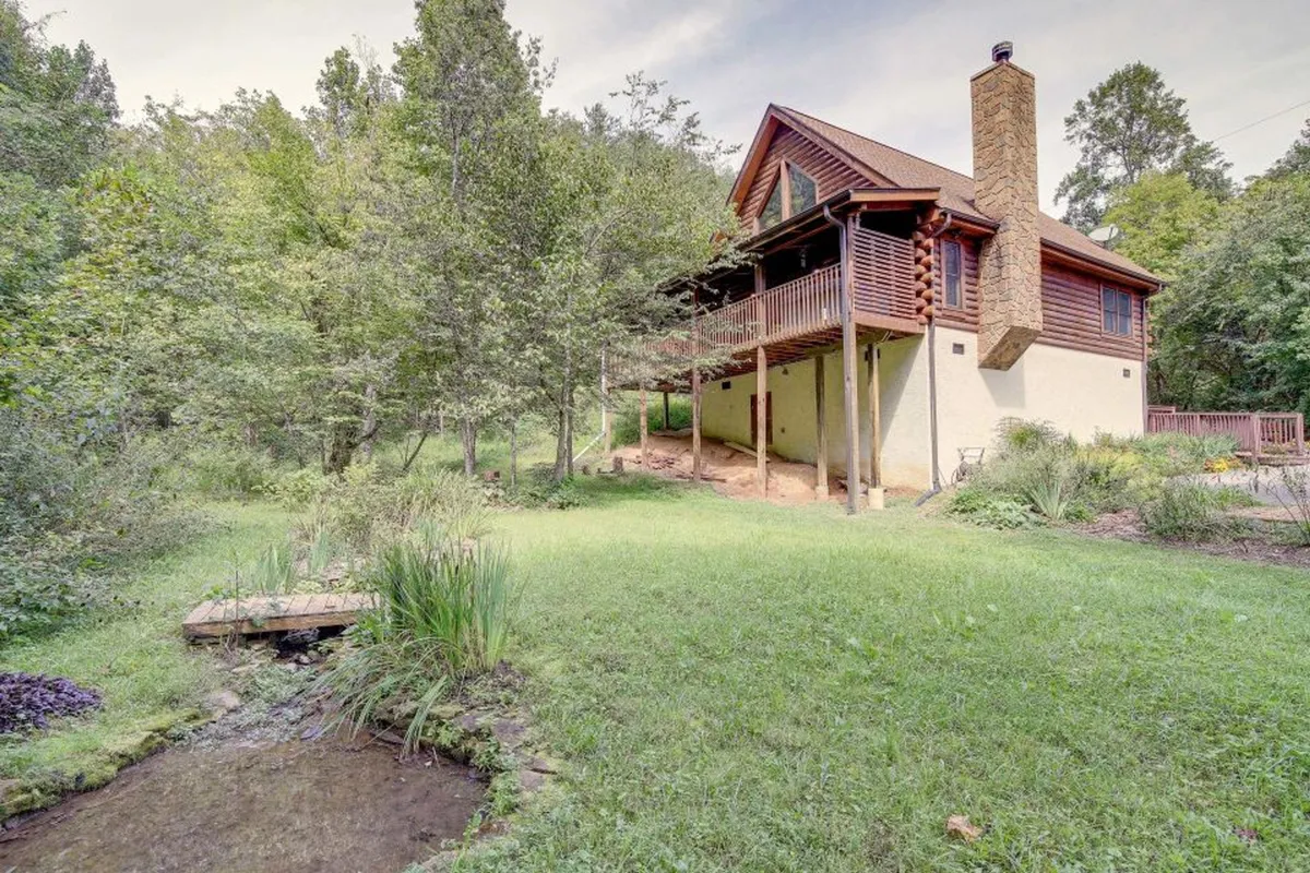 The image showcases a charming, newly-renovated cabin nestled amid tall, leafy trees on a private, landscaped lot. A serene creek runs beside the property, enhancing the sense of seclusion and natural beauty. The inviting front porch features a swing, perfect for relaxing and enjoying the tranquil sounds of nature. Lush greenery surrounds the scene, creating an atmosphere of peaceful retreat.