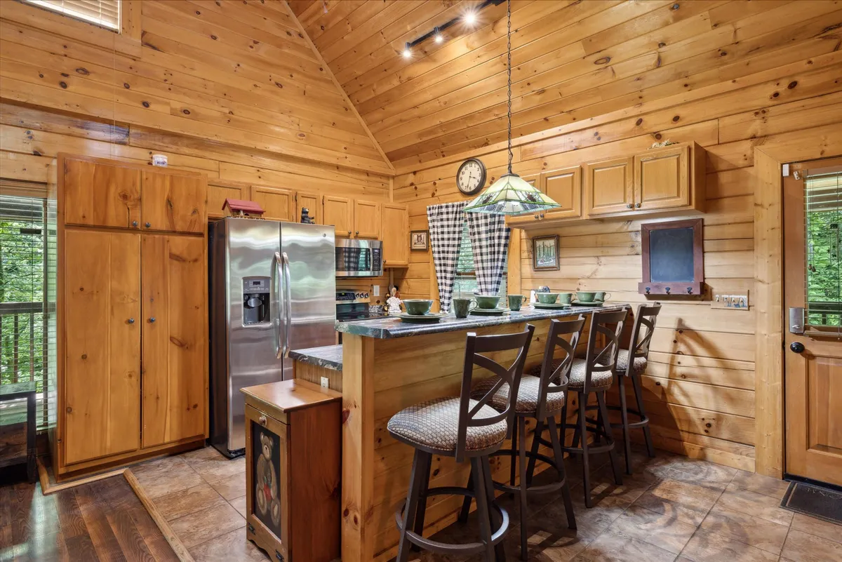 This image centers on a welcoming kitchen space featuring wooden cabinetry, a stainless steel refrigerator, bar seating, and a stylish hanging light fixture. The kitchen is open and inviting, with space for multiple cooks or guests to gather and enjoy the ambiance. The view from the kitchen includes nature outside, adding a tranquil touch.