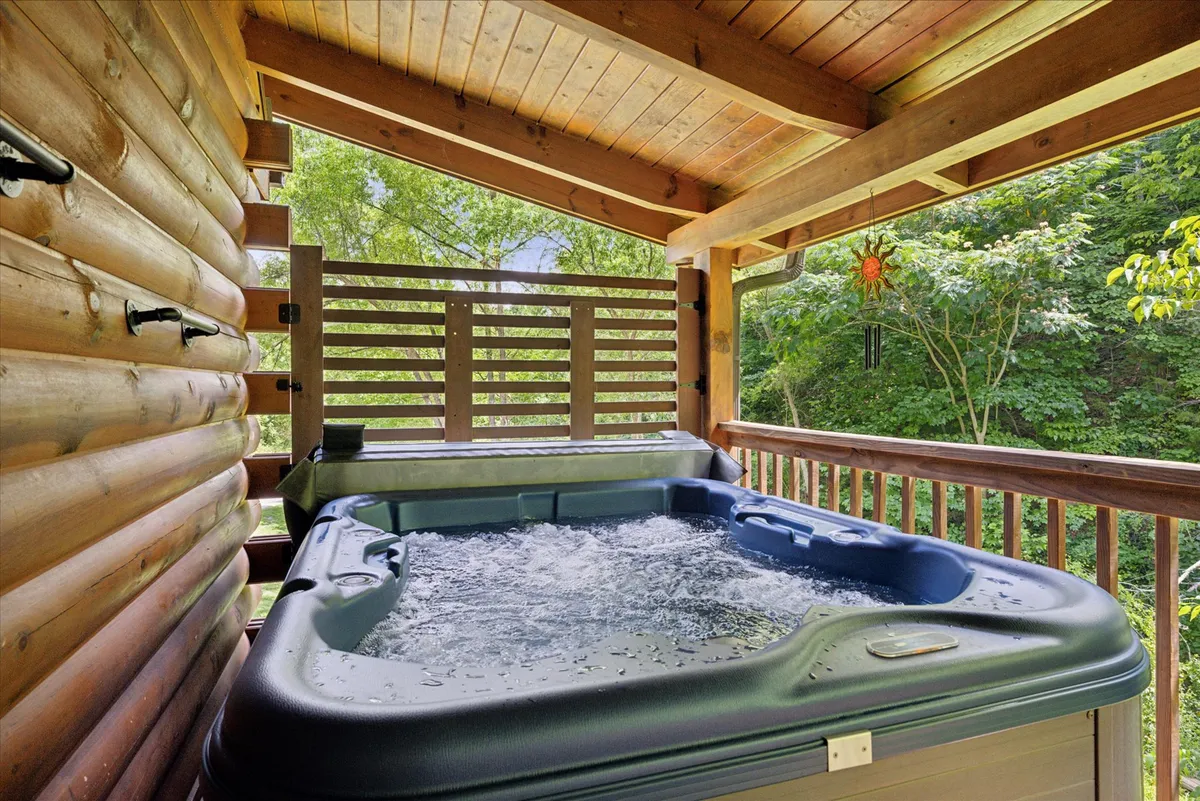 A private hot tub sits on a wooden deck, surrounded by lush greenery and a simple wooden railing. The hot tub appears inviting and secluded, ideal for unwinding after outdoor adventures. Natural sunlight filters through the trees, enhancing the sense of privacy and calm.