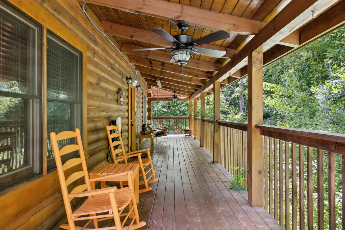 The image shows a cozy porch with classic wooden rocking chairs lined up along a sturdy wooden railing. Overhead fans spin lazily, and the wide porch looks out onto a peaceful natural setting with trees visible in the background. The setting exudes tranquility and invites relaxation or conversation while taking in the sights and sounds of nature.