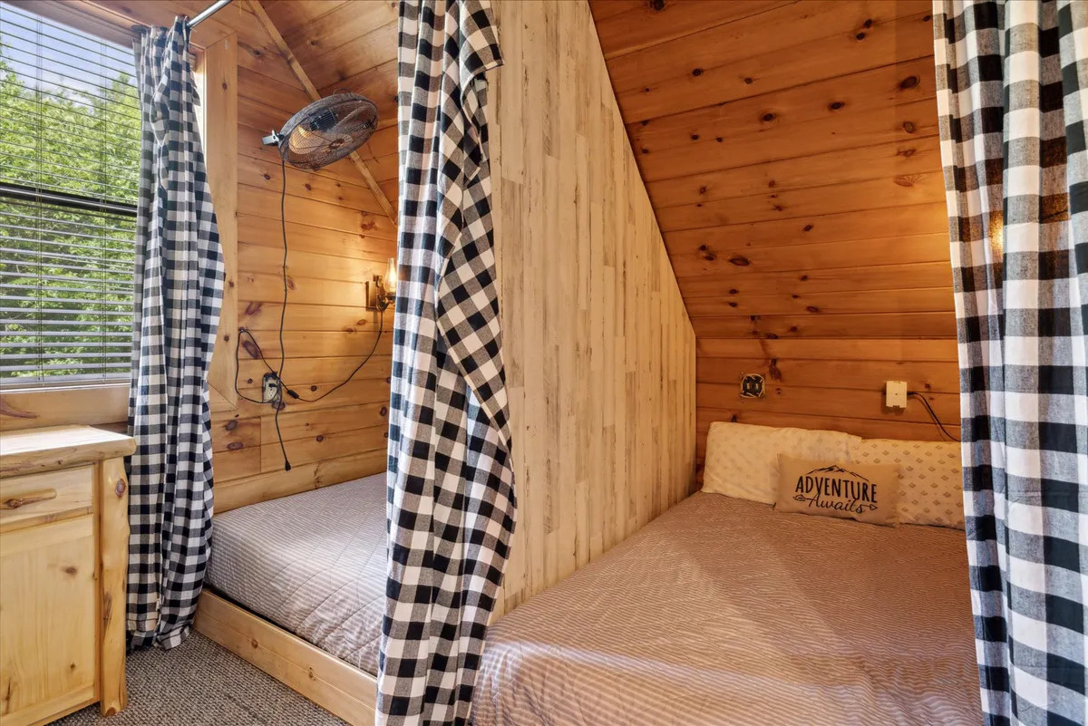 A cozy bedroom with two beds featuring patterned bedding, wooden walls, and black and white checkered curtains. A decorative pillow with text adds a playful touch, evoking warmth and togetherness.