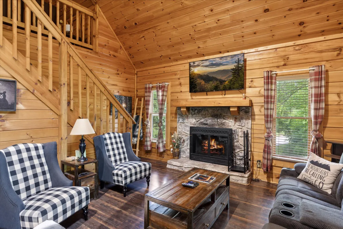 A spacious living space anchored by a prominent fireplace, with a plush sofa and checkered chairs arranged for socializing or game nights. The wooden staircase in the background adds rustic charm, and the area feels ready for family gatherings or relaxed evenings.